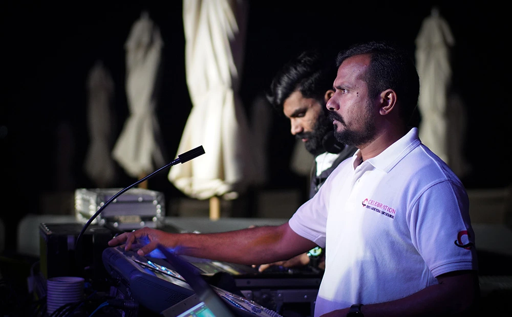 Two men operate a DJ mixer at night, with a microphone in front and white polo in the foreground.