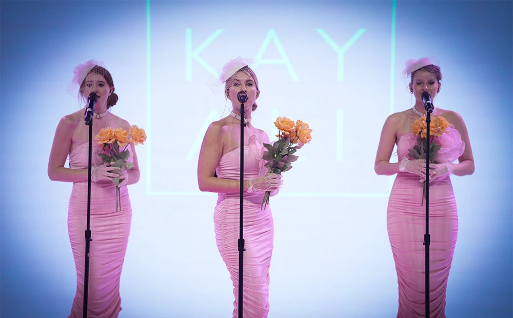 Three female singers in pink gowns perform on stage, each holding yellow roses and singing into a microphone against a blue-lit backdrop.