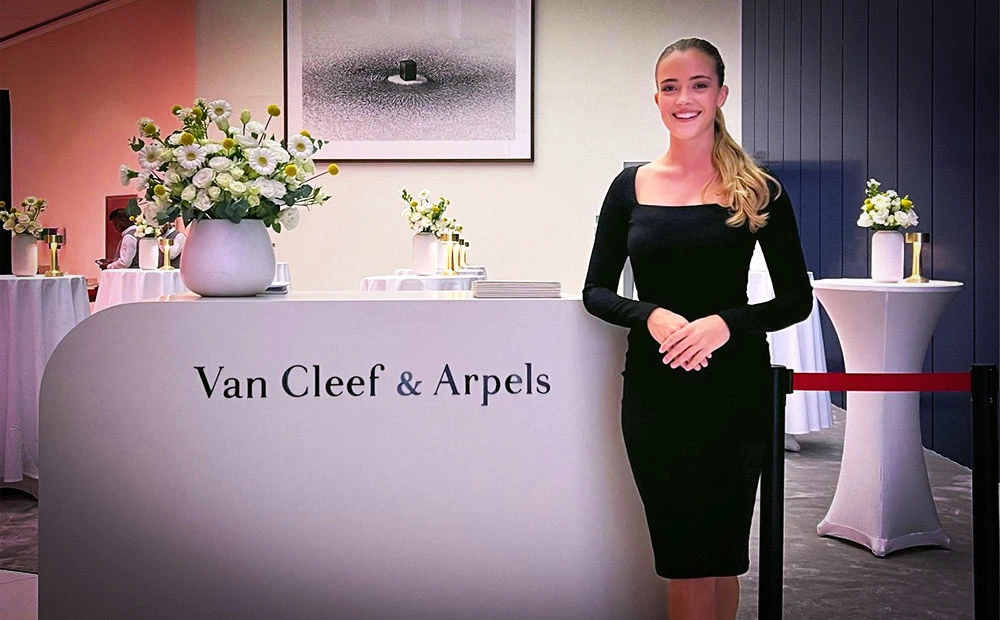 Smiling woman in a black dress stands at a Van Cleef & Arpels reception desk with a white floral centerpiece nearby.
