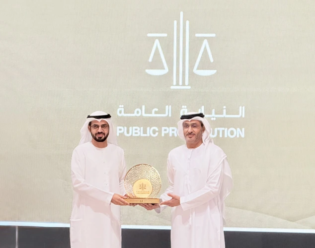 Two men in white kanduras present a gold circular award on a stage, with the Public Prosecution logo and Arabic text in the background.