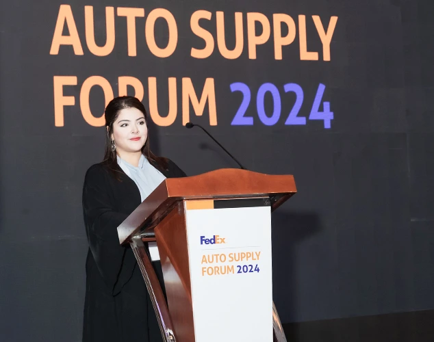 Woman in professional attire stands at a podium on stage, with a large 'Auto Supply Forum 2024' backdrop and a FedEx-branded podium sign.